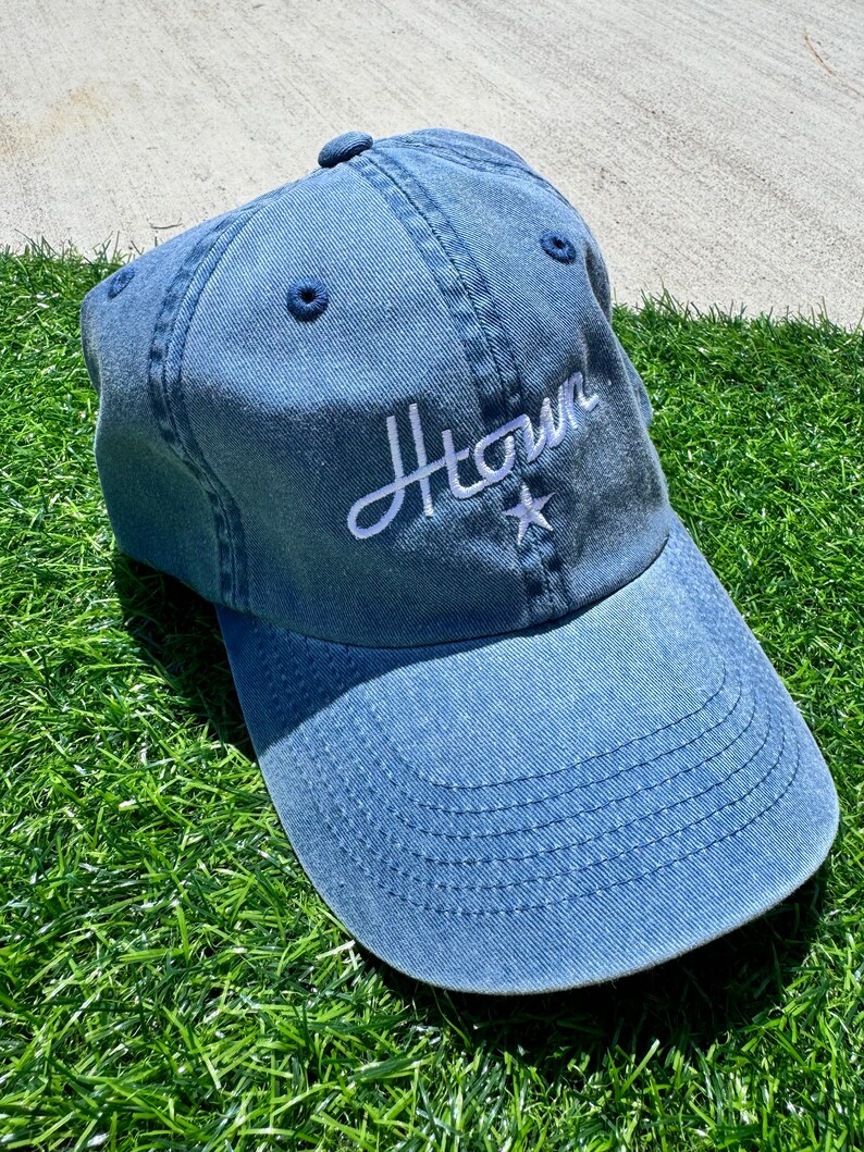 May include: A denim blue baseball cap with the word "Htown" embroidered in white script. A white star is embroidered below the word. The cap is sitting on a bed of green grass.