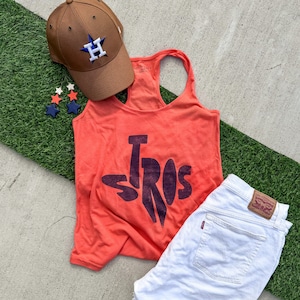 May include: An orange tank top with the word "STROS" in dark purple, a brown baseball cap with a white and blue logo, white denim shorts, and star-shaped earrings. The items are arranged on a concrete surface with a strip of artificial turf.