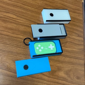 Peut inclure: Plusieurs petits &eacute;tuis rectangulaires de diff&eacute;rentes couleurs sont pr&eacute;sent&eacute;s. L'un est bleu clair, un autre gris et un troisi&egrave;me blanc. Une manette de jeu verte avec des boutons blancs et une croix est &agrave; l'int&eacute;rieur d'un &eacute;tui bleu. Chaque &eacute;tui a un point noir.