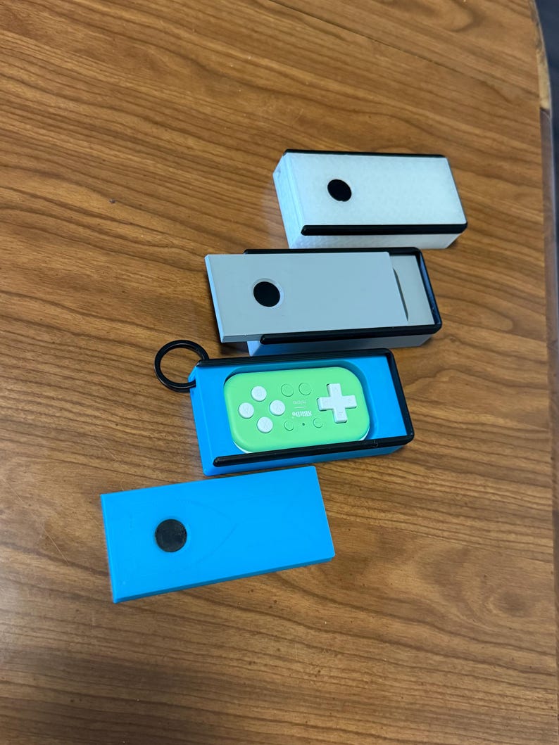 Peut inclure: Plusieurs petits &eacute;tuis rectangulaires de diff&eacute;rentes couleurs sont pr&eacute;sent&eacute;s. L'un est bleu clair, un autre gris et un troisi&egrave;me blanc. Une manette de jeu verte avec des boutons blancs et une croix est &agrave; l'int&eacute;rieur d'un &eacute;tui bleu. Chaque &eacute;tui a un point noir.