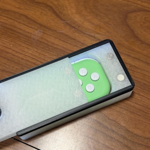 Peut inclure: Un &eacute;tui rectangulaire blanc et noir avec une manette verte &agrave; l'int&eacute;rieur. La manette a des boutons blancs. L'&eacute;tui est partiellement ouvert, r&eacute;v&eacute;lant la manette. Le fond est une surface en bois.