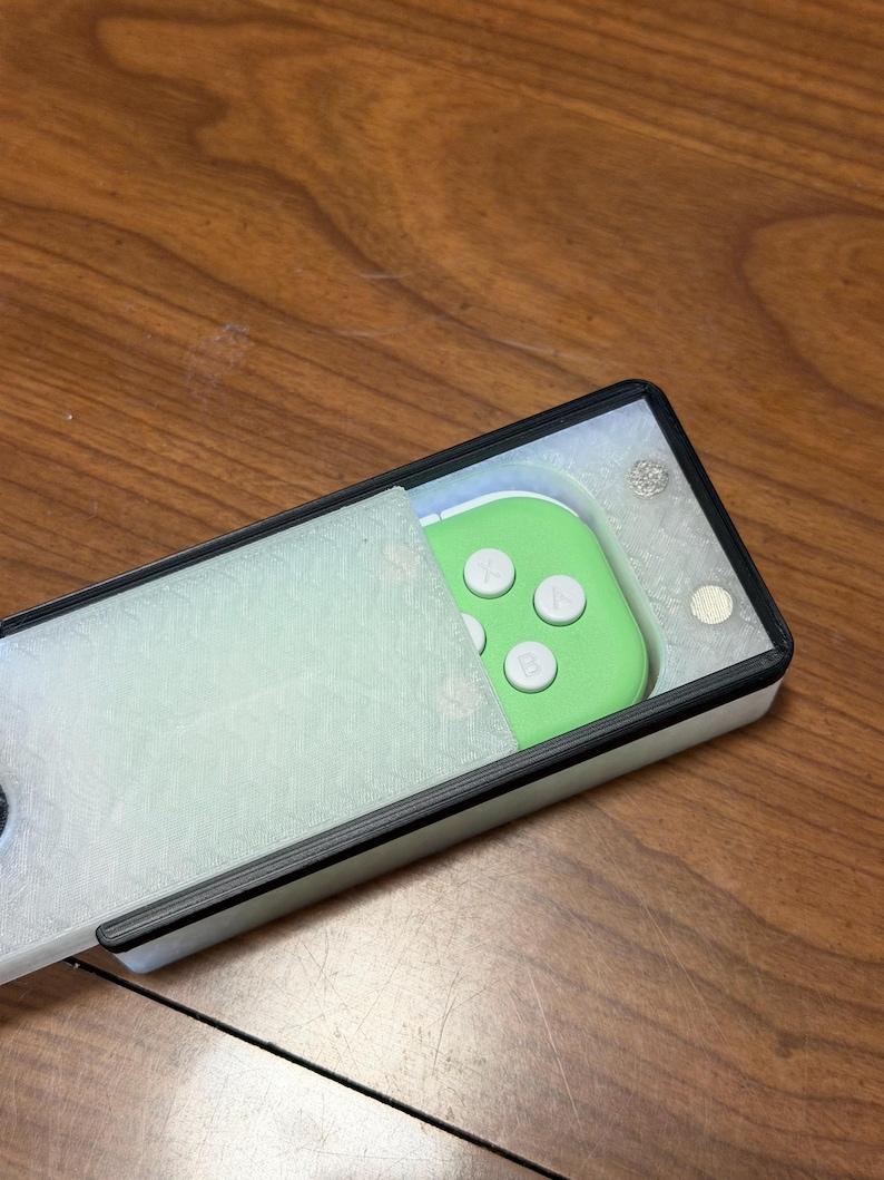 Peut inclure: Un &eacute;tui rectangulaire blanc et noir avec une manette verte &agrave; l'int&eacute;rieur. La manette a des boutons blancs. L'&eacute;tui est partiellement ouvert, r&eacute;v&eacute;lant la manette. Le fond est une surface en bois.