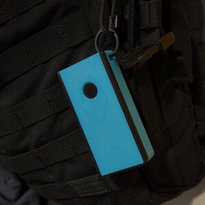 Peut inclure: Un objet rectangulaire bleu clair avec une bordure noire et un trou circulaire noir, attach&eacute; &agrave; un sac &agrave; dos noir via un mousqueton. L'objet est de couleur unie et semble &ecirc;tre en plastique.