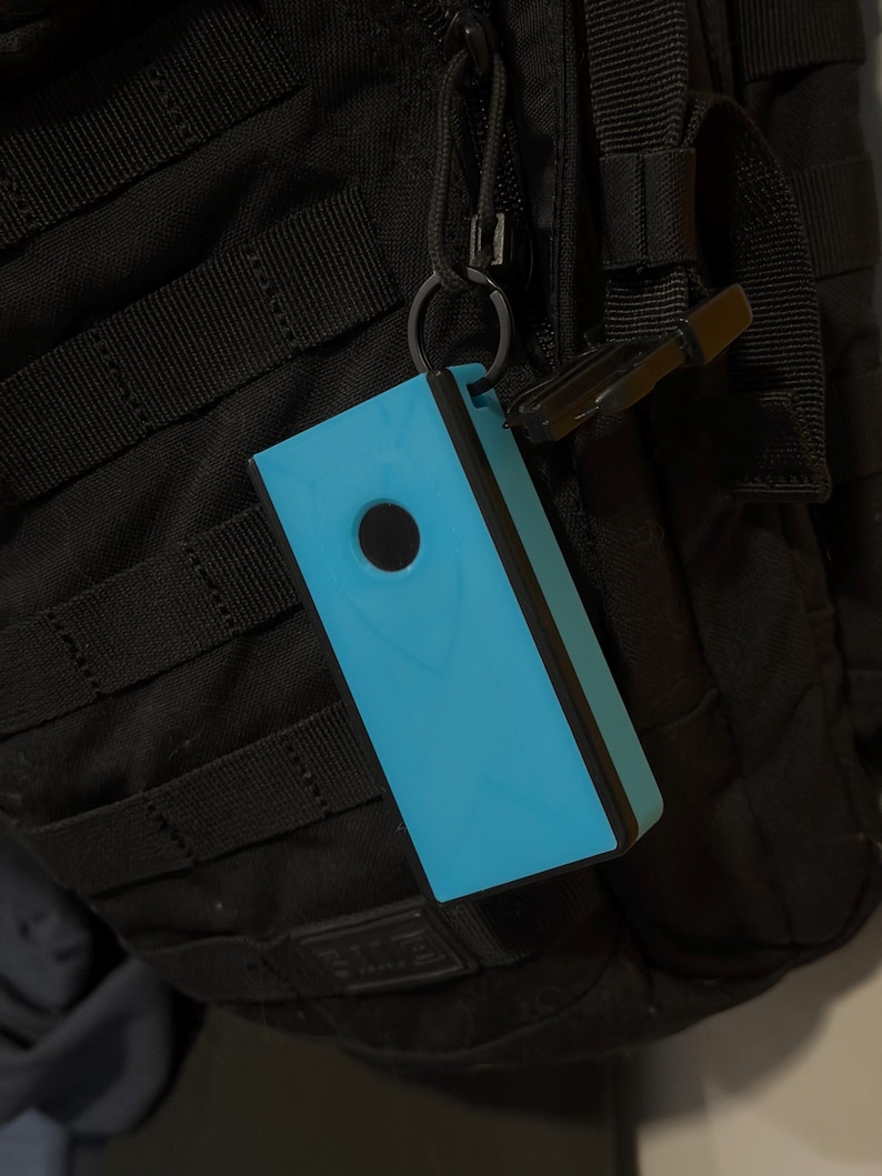 Peut inclure: Un objet rectangulaire bleu clair avec une bordure noire et un trou circulaire noir, attach&eacute; &agrave; un sac &agrave; dos noir via un mousqueton. L'objet est de couleur unie et semble &ecirc;tre en plastique.