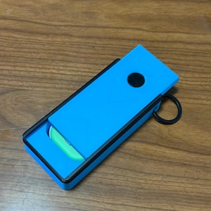 Peut inclure: Un &eacute;tui rectangulaire bleu avec une bordure noire et un bouton circulaire noir. L'&eacute;tui est ouvert, r&eacute;v&eacute;lant un objet vert &agrave; l'int&eacute;rieur. Un anneau noir est attach&eacute; sur le c&ocirc;t&eacute;. L'&eacute;tui est sur une surface en bois.