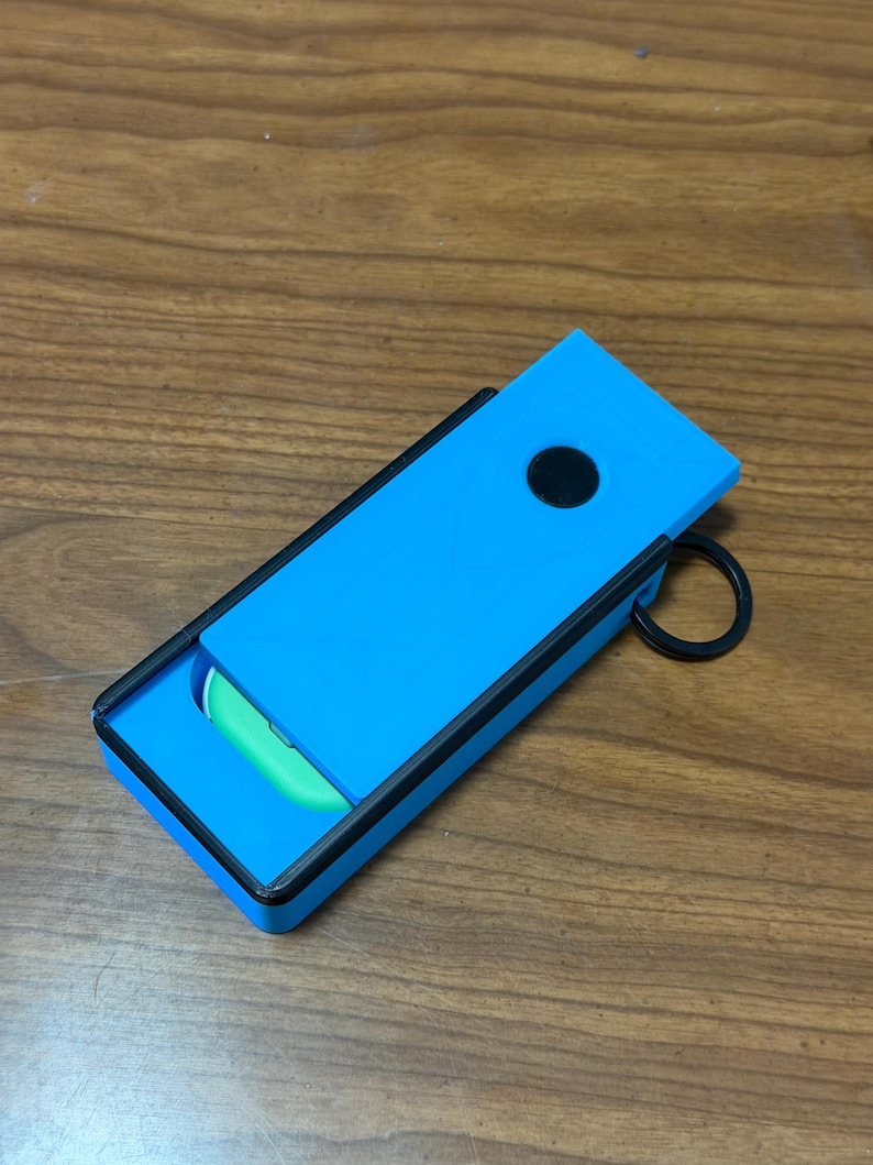 Peut inclure: Un &eacute;tui rectangulaire bleu avec une bordure noire et un bouton circulaire noir. L'&eacute;tui est ouvert, r&eacute;v&eacute;lant un objet vert &agrave; l'int&eacute;rieur. Un anneau noir est attach&eacute; sur le c&ocirc;t&eacute;. L'&eacute;tui est sur une surface en bois.