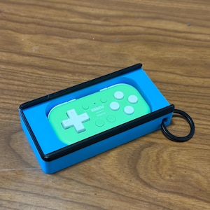 Peut inclure: Une manette de jeu portable verte et blanche, &eacute;tiquet&eacute;e "8BitDo micro", &agrave; l'int&eacute;rieur d'un &eacute;tui bleu et noir. La manette poss&egrave;de une croix directionnelle et des boutons. Un anneau noir est attach&eacute; &agrave; l'&eacute;tui.
