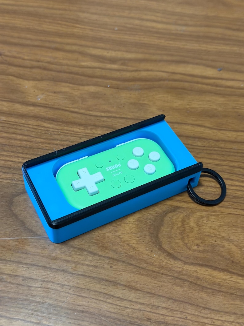 Peut inclure: Une manette de jeu portable verte et blanche, &eacute;tiquet&eacute;e "8BitDo micro", &agrave; l'int&eacute;rieur d'un &eacute;tui bleu et noir. La manette poss&egrave;de une croix directionnelle et des boutons. Un anneau noir est attach&eacute; &agrave; l'&eacute;tui.