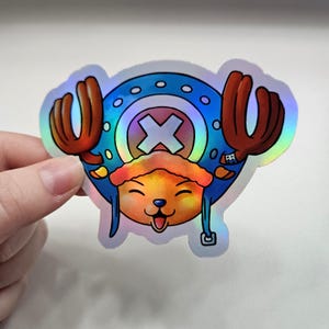 Puede incluir: Pegatina holográfica con un personaje de dibujos animados con un sombrero azul y cuernos, un símbolo de cruz y una cara sonriente. La pegatina tiene un acabado brillante e iridiscente, que refleja varios colores. El personaje tiene una expresión feliz con la lengua fuera.