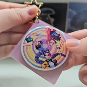 以下が含まれることがあります： 長い髪と武器を持った3人のキャラクターの漫画イラストが描かれた、丸くてカラフルなキーホルダー。キーホルダーには金色の星型の留め金と、薄紫色の裏地が付いています。