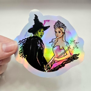 May include: Holographic sticker featuring illustrations of two characters from the musical "Wicked". One character is a green-skinned witch in a black hat and cape. The other is a blonde woman in a pink dress and crown, holding a wand.