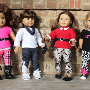 May include: Four dolls wearing different outfits. The dolls are wearing pink, red, white, and black shirts. The dolls are wearing patterned leggings. The dolls are standing in front of a brick wall.