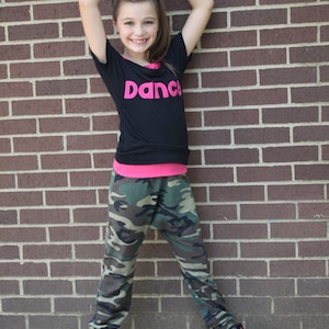 May include: A young person wearing a black shirt with the word "Dance" in pink letters, camouflage trousers, and black and pink trainers. The person is standing in front of a brick wall.