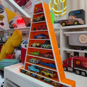 May include: An orange and white display shelf shaped like a motel sign, filled with toy cars. The shelf has the words "COZY CONE MOTEL" on the side. The cars are various colours and styles. The shelf is on a white surface.
