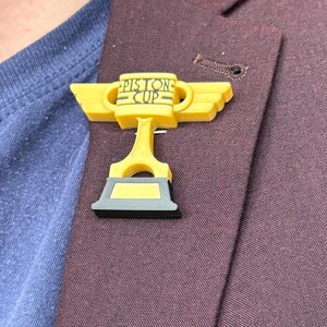 Puede incluir: Un pin de trofeo Piston Cup amarillo con detalles en negro y gris. El pin est&aacute; sujeto a una blazer granate. El texto "Piston Cup" es visible en el trofeo.