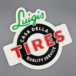 May include: A white, rectangular sticker with a black and red logo. The logo features the words "Luigis" in green script above a black circle with "CASA DELLA TIRES QUALITY SERVICE" and the word "TIRES" in red.