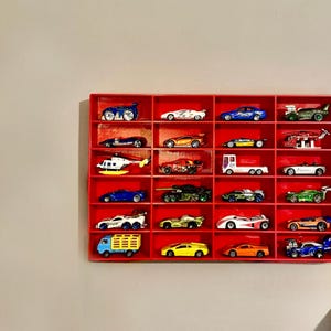 A 3D-printed red wall-mounted display shelf holding 24 toy cars, including sports cars, trucks, and a helicopter. The compact storage solution keeps diecast vehicles neatly organised and easily accessible.