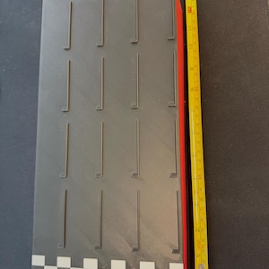 May include: A rectangular, gray object with vertical slots and a red side. The object has a checkered pattern on one end. A yellow tape measure shows the object is approximately 13 inches long.