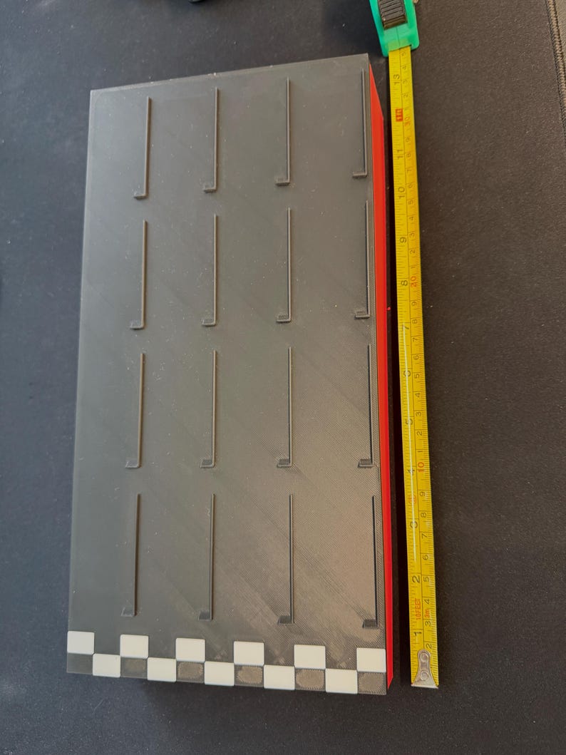 May include: A rectangular, gray object with vertical slots and a red side. The object has a checkered pattern on one end. A yellow tape measure shows the object is approximately 13 inches long.