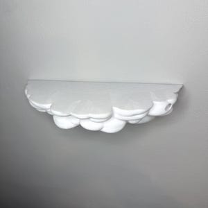 May include: A white, cloud-shaped wall shelf. The shelf has a flat top and a scalloped, cloud-like bottom edge. The shelf is made of a solid material and is mounted on a white wall. The shelf is designed to hold small items.