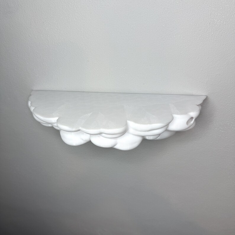 May include: A white, cloud-shaped wall shelf. The shelf has a flat top and a scalloped, cloud-like bottom edge. The shelf is made of a solid material and is mounted on a white wall. The shelf is designed to hold small items.