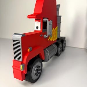 May include: A red toy semi-truck with a silver grill, wheels, and exhaust pipes. The truck has a tall cab and a yellow flame design on the side. The truck is a toy.