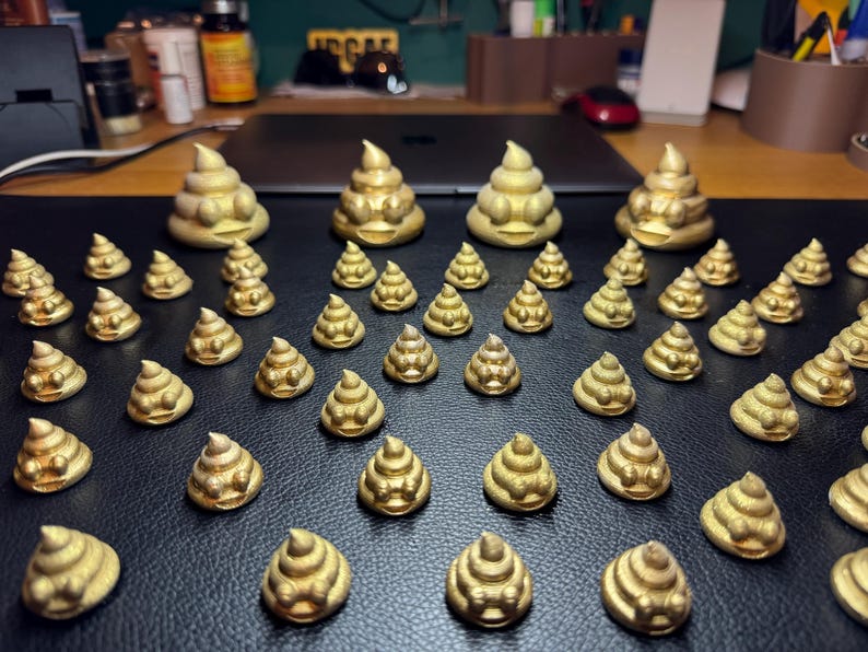A mixture of 5cm and 2cm tall golden glittered poop emoji 3D prints rest on a black leather surface. All of the poops are smiling with eyes slightly protruding from their heads.