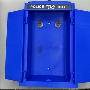 May include: A blue Police Box with the words "POLICE PUBLIC CALL BOX" in white lettering. The box has two doors that are open, revealing the interior. The box has two silver-colored knobs inside.