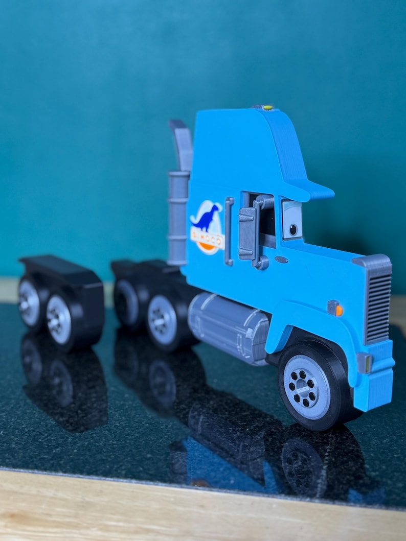 May include: A light blue toy semi-truck with a cartoonish design. The truck features a cab with a visor, a trailer, and a logo with a blue dinosaur. The wheels are gray and black.