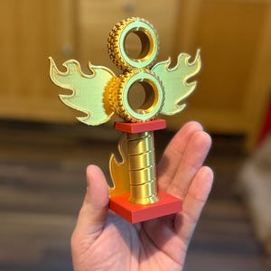 May include: A gold-coloured trophy with a red base, featuring two stacked tyre-shaped elements and stylized flame and wing accents. The trophy is held in a hand, showcasing its size and design.