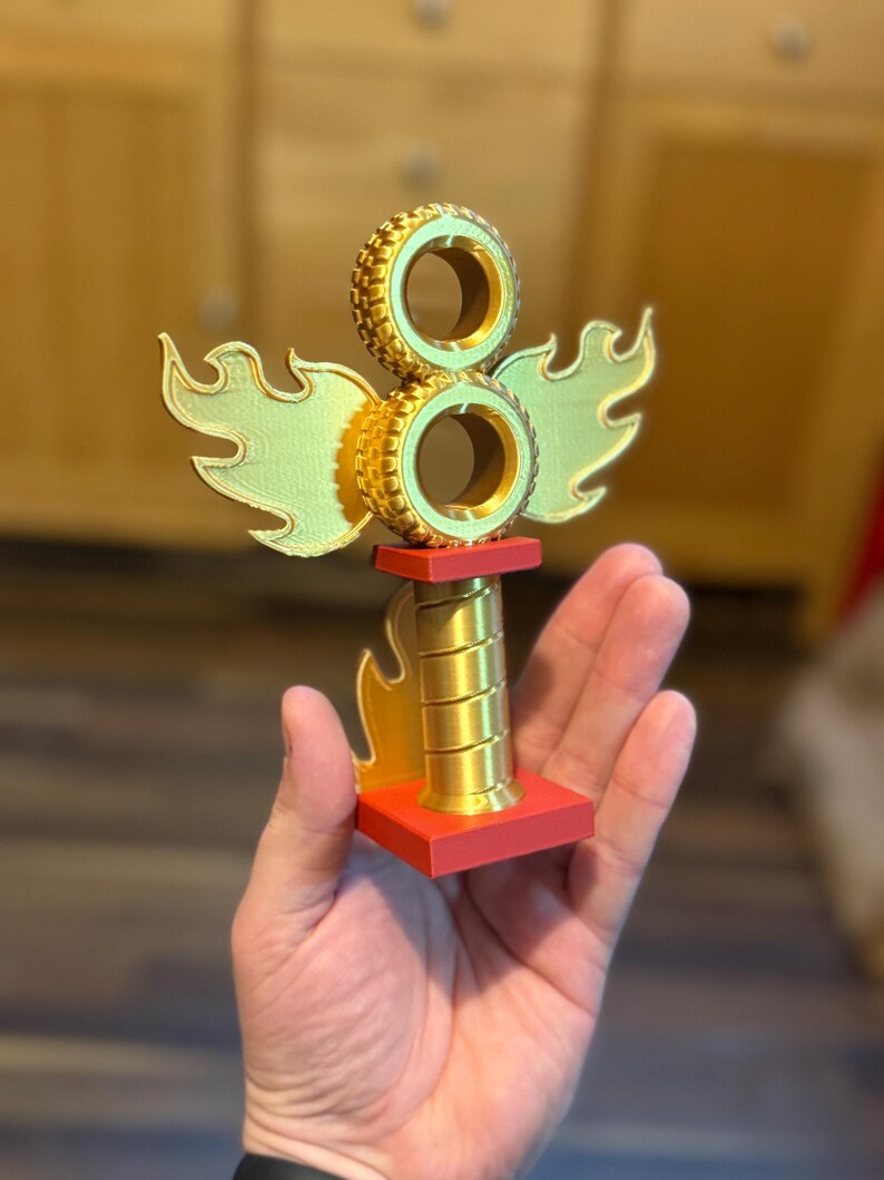 May include: A gold-coloured trophy with a red base, featuring two stacked tyre-shaped elements and stylized flame and wing accents. The trophy is held in a hand, showcasing its size and design.