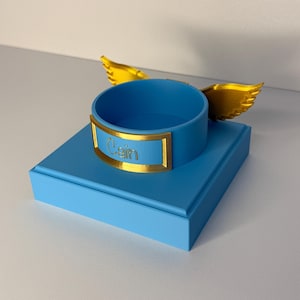 May include: A light blue square base supports a circular blue ring holder featuring a gold-colored nameplate with the text "Cain" embossed in the center. Two gold-colored wings extend from the sides of the circular holder, adding detail.