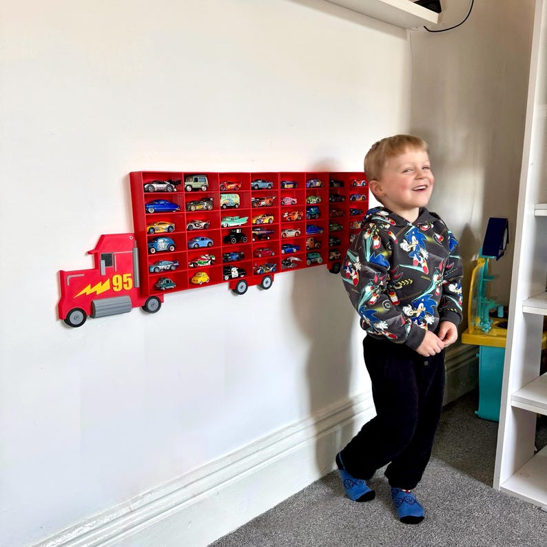Happy kid stood next to his birthday present gift that is a Red Disney-inspired toy car storage truck with shelving, perfect for holding Hot Wheels or Matchbox cars. This fun practical display is a great way to encourage your child to tidy their toys