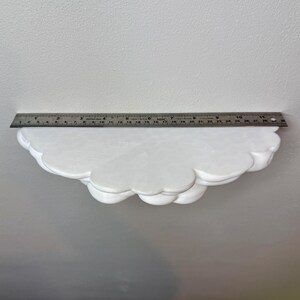 May include: A white cloud-shaped shelf with a scalloped edge. A metal ruler is placed on top of the shelf, measuring approximately 11 inches in length. The shelf is made of a solid material and is mounted on a white wall.
