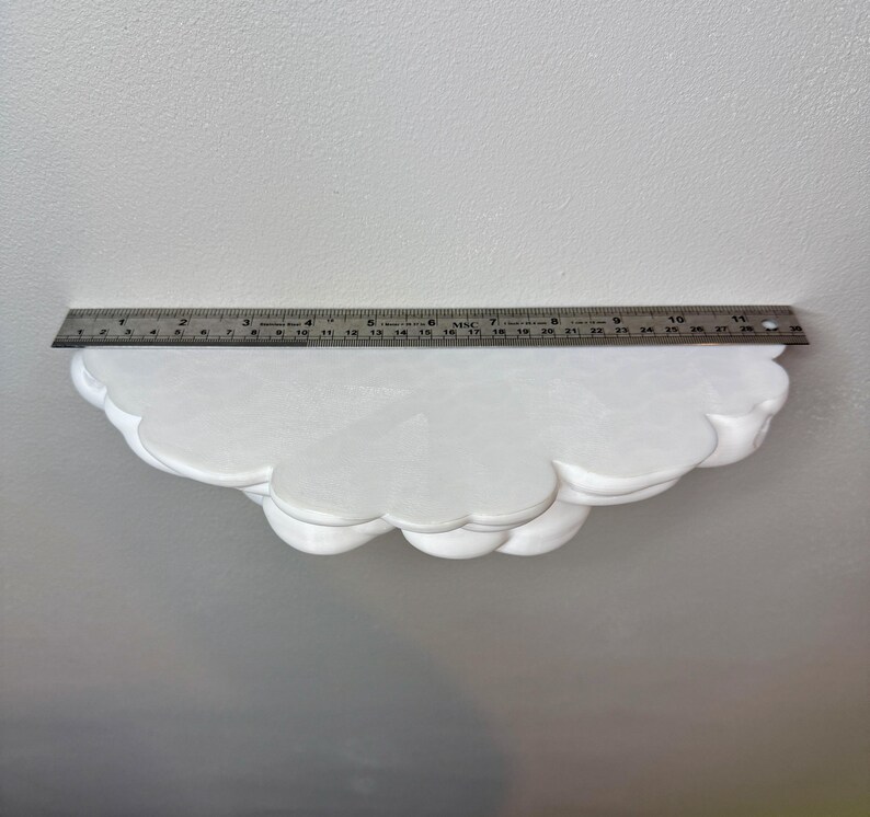 May include: A white cloud-shaped shelf with a scalloped edge. A metal ruler is placed on top of the shelf, measuring approximately 11 inches in length. The shelf is made of a solid material and is mounted on a white wall.