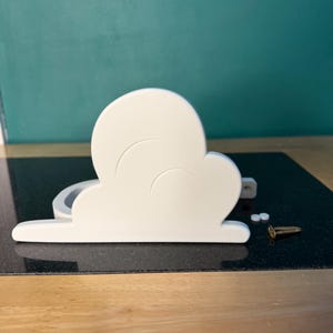 May include: A white cloud-shaped decorative object with a curved design. The object is made of a solid material and has a flat base. Two small white pieces and a gold screw are next to the cloud. The background is a teal wall.