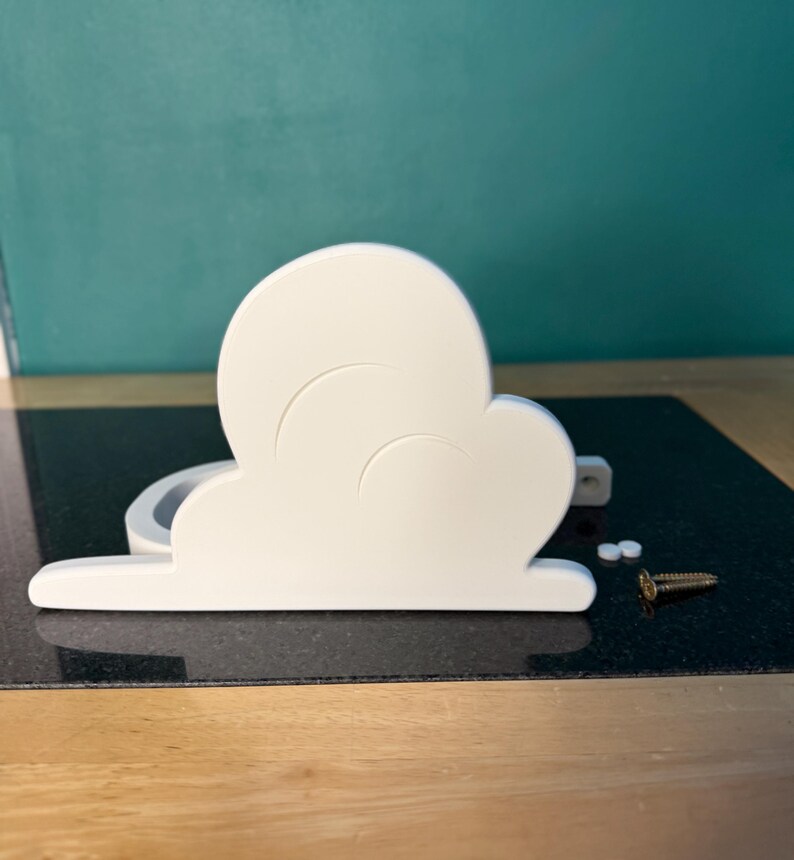 May include: A white cloud-shaped decorative object with a curved design. The object is made of a solid material and has a flat base. Two small white pieces and a gold screw are next to the cloud. The background is a teal wall.