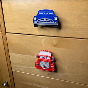 May include: Two toy car drawer pulls. The top pull is a blue car with white accents. The bottom pull is a red truck with a white roof. Both pulls have cartoon-like eyes and are mounted on a wooden drawer.