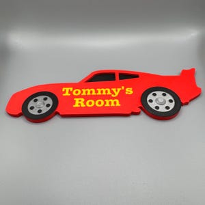May include: A red car-shaped sign with the text "Tommy's Room" in yellow. The sign has black and silver wheels and a spoiler. The sign is designed to be mounted on a wall.