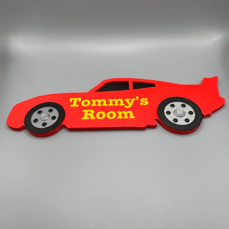 May include: A red car-shaped sign with the text "Tommy's Room" in yellow. The sign has black and silver wheels and a spoiler. The sign is designed to be mounted on a wall.