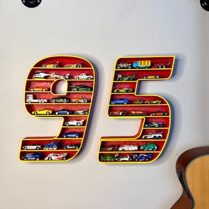 May include: A wall-mounted display shelf shaped like the number 95, painted yellow with a red interior. The shelves are filled with miniature toy cars of various colors and designs. The display is mounted on a white wall.