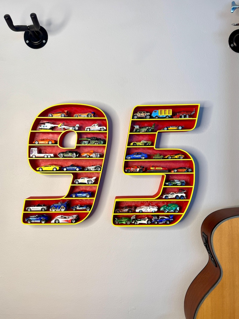 May include: A wall-mounted display shelf shaped like the number 95, painted yellow with a red interior. The shelves are filled with miniature toy cars of various colors and designs. The display is mounted on a white wall.
