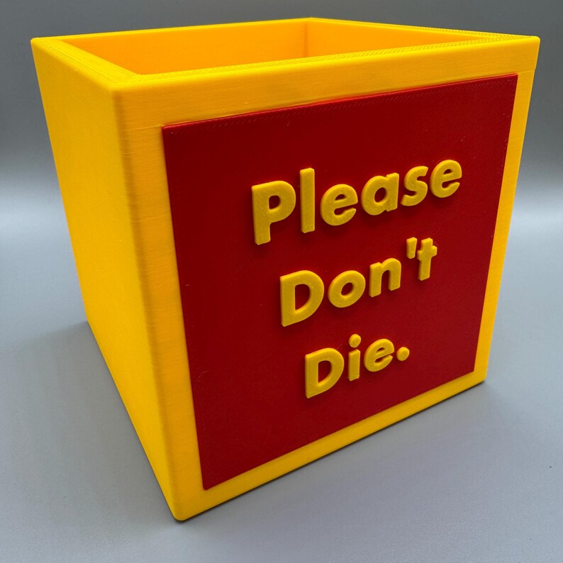 May include: A yellow square container with a red rectangular panel. The panel features the phrase "Please Don't Die" in yellow, raised lettering. The container is likely 3D-printed.