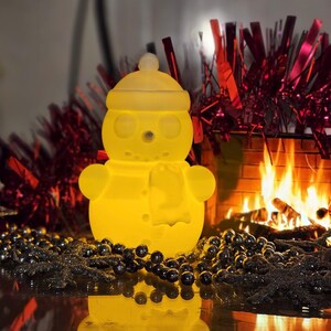 Yellow glowing 3D-printed snowman LED tealight holder, displayed on a festive table with red tinsel, silver beads, and a cosy fireplace in the background.