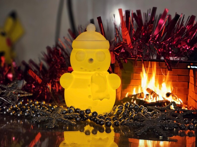 Yellow glowing 3D-printed snowman LED tealight holder, displayed on a festive table with red tinsel, silver beads, and a cosy fireplace in the background.