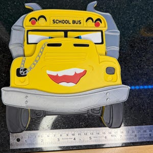 K&ouml;nnte beinhalten: Gelbe, schulbusf&ouml;rmige Dekoration mit einem l&auml;chelnden Gesicht, rotem Mund und dem Schriftzug "SCHOOL BUS" oben. Der Bus hat graue Akzente und ein Kettendetail. Das Bild enth&auml;lt ein Lineal zur Ma&szlig;stabsbestimmung.