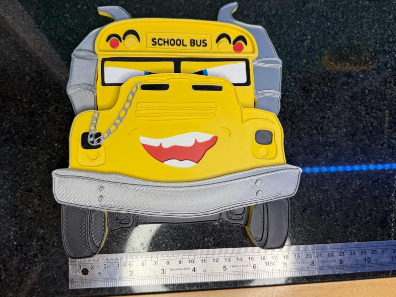 K&ouml;nnte beinhalten: Gelbe, schulbusf&ouml;rmige Dekoration mit einem l&auml;chelnden Gesicht, rotem Mund und dem Schriftzug "SCHOOL BUS" oben. Der Bus hat graue Akzente und ein Kettendetail. Das Bild enth&auml;lt ein Lineal zur Ma&szlig;stabsbestimmung.