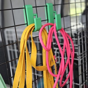 Close-up of two green 3D-printed hooks attached to a dog crate, holding yellow and pink longlines. The hooks clip securely over the metal bars, keeping the leads neatly organised in a vehicle setup.