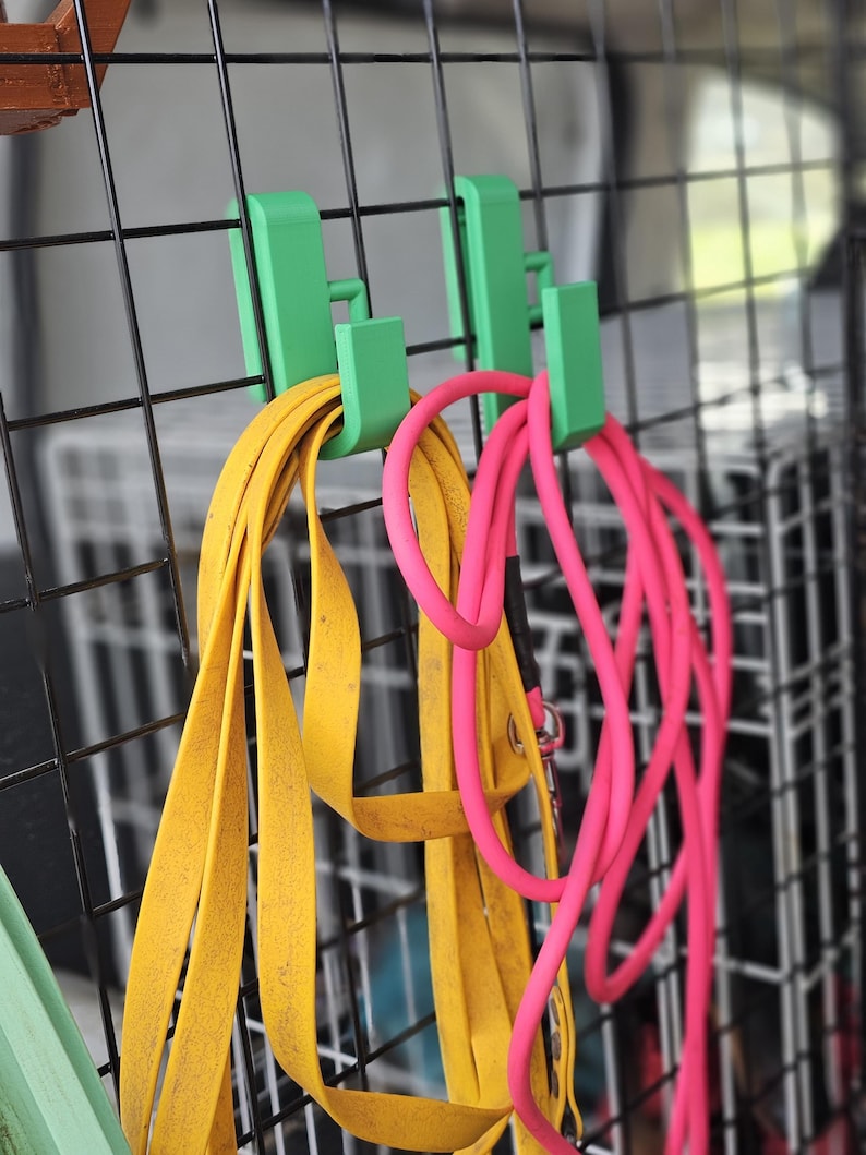 Close-up of two green 3D-printed hooks attached to a dog crate, holding yellow and pink longlines. The hooks clip securely over the metal bars, keeping the leads neatly organised in a vehicle setup.