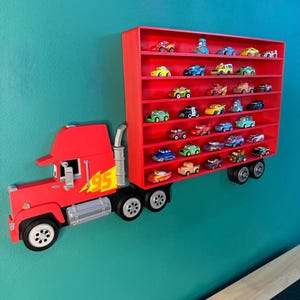 May include: A red wall-mounted display shelf shaped like a semi-truck trailer, featuring multiple horizontal tiers filled with various small toy cars. The truck cab is red with a yellow number 95 lightning bolt graphic and silver exhaust pipes.